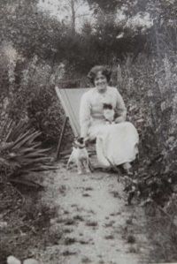 Photo of Hilda Burkitt sitting on a deck chair in a garden. She is wearing a white dress and has dark curly hair.  She's stroking a small dog that's standing beside her.